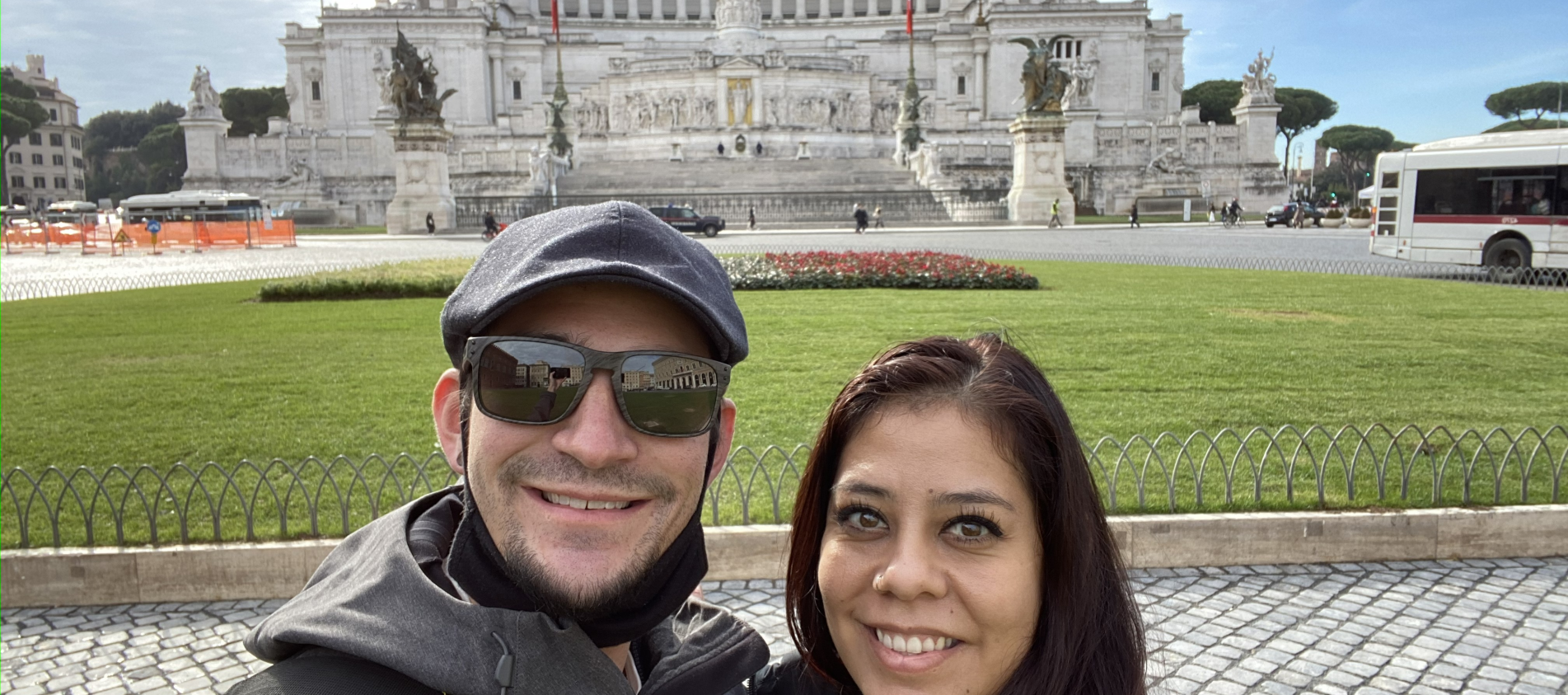 Couple smiling in front of the Altare della Patria monument in Rome, Italy, on a sunny day.