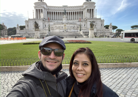 Couple smiling in front of the Altare della Patria monument in Rome, Italy, on a sunny day.